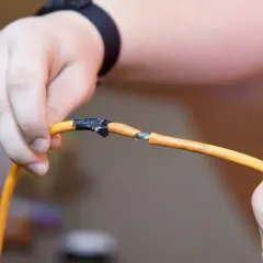 Dłonie trzymają przecięty pomarańczowy kabel elektryczny, pokazując, jak połączyć przecięty kabel elektryczny przy użyciu taśmy izolacyjnej.