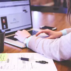 Kobieta pracuje przy laptopie. Na biurku leżą notatki, długopis i kubek. Dobre natężenie oświetlenia na stanowisku pracy sprzyja koncentracji.
