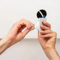 Dłonie montują oprawę lampy sufitowej, przygotowując ją do podłączenia. Widać tu fragmenty przewodów i otwór w suficie, co sugeruje instalację. Pytanie, jakie kołki do lampy sufitowej, jest kluczowe dla stabilnego montażu.