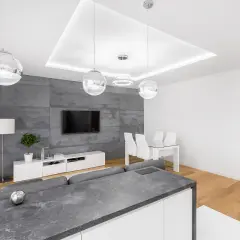 Nowoczesny salon z szarą ścianą, białym meblem RTV i wnęką na led w suficie. W tle stół z krzesłami.