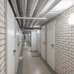 Korytarz piwnicy z rzędami drzwi i rurami na suficie. Oświetlenie piwnicy zapewnia jasne, okrągłe lampy.