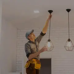 Fachowiec w pasie z narzędziami dokonuje montażu kinkietu, wiszącego obok innych lamp.