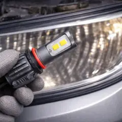 Ręka w rękawiczce trzyma żarówkę LED przed reflektorem samochodowym.