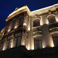 Elegancka kamienica nocą, oświetlona ciepłym blaskiem lamp miejskich. Widać bogate zdobienia fasady i duże, łukowe okna.