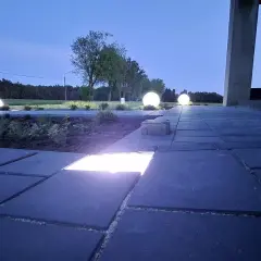 Nocny ogród rozświetlony kulistymi lampami i ledami w kostce brukowej. Ścieżka z ciemnej kostki brukowej prowadzi do domu.