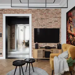 Nowoczesny salon z ceglaną ścianą, na której wisi telewizor. Oświetlenie ściany z kamienia podkreśla jej fakturę. Obok żółty fotel i obraz z gitarzystą.
