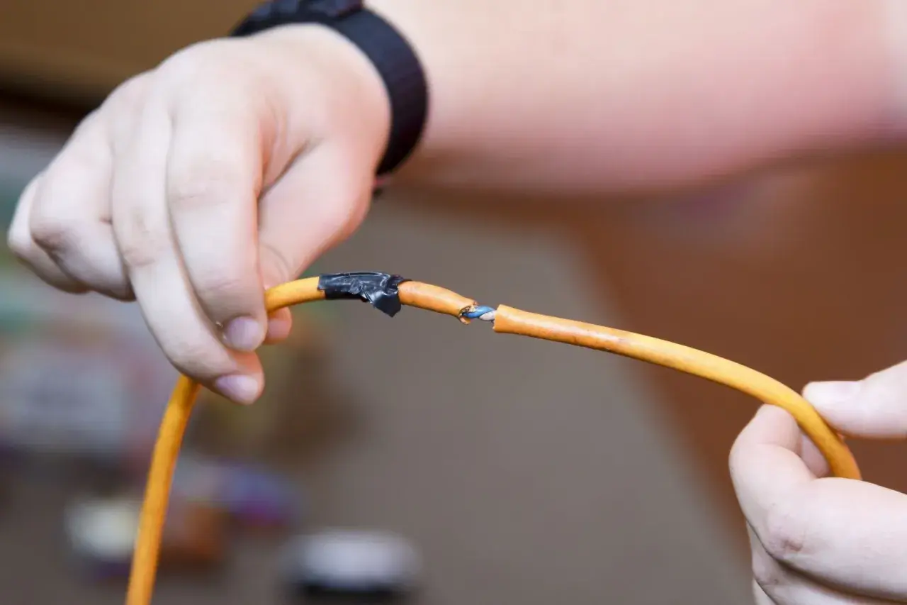 Dłonie trzymają przecięty pomarańczowy kabel elektryczny, pokazując, jak połączyć przecięty kabel elektryczny przy użyciu taśmy izolacyjnej.