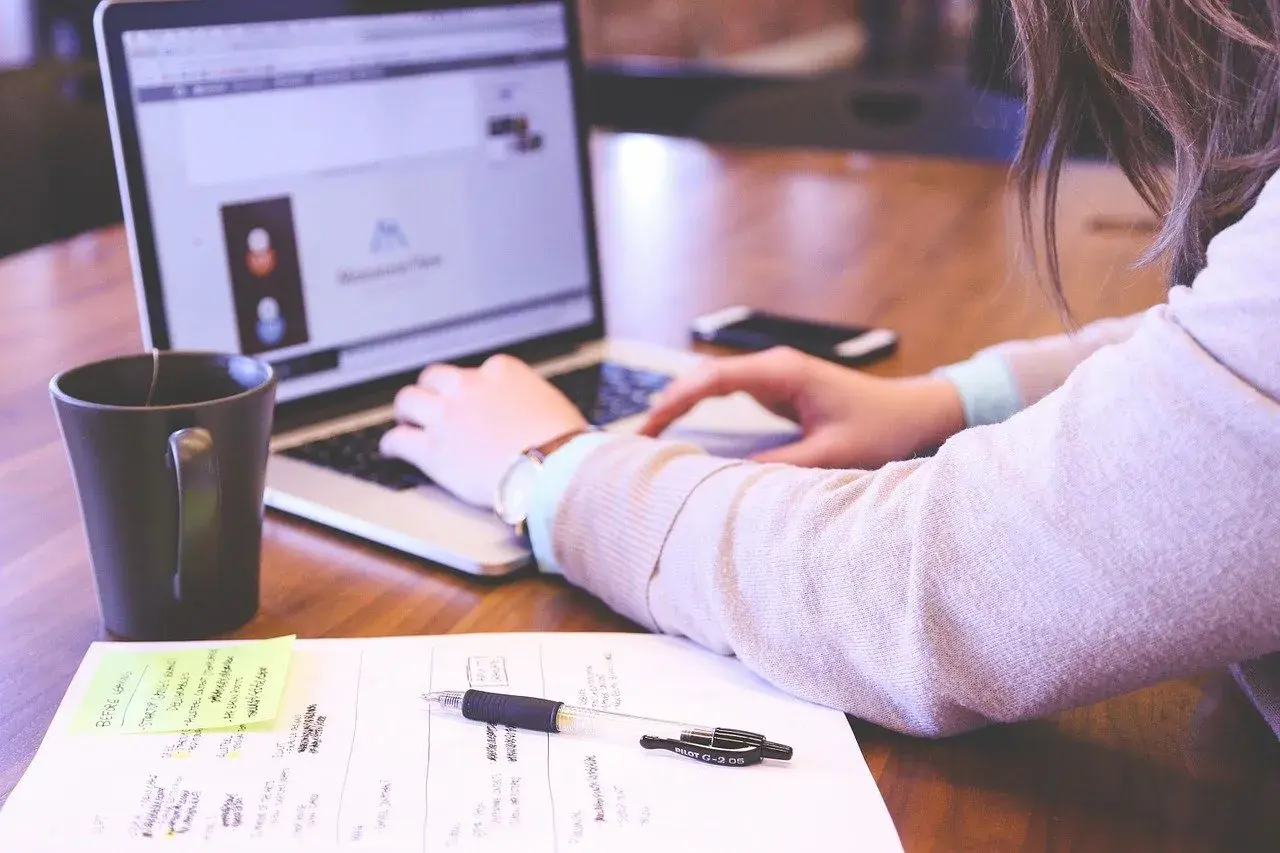 Kobieta pracuje przy laptopie. Na biurku leżą notatki, długopis i kubek. Dobre natężenie oświetlenia na stanowisku pracy sprzyja koncentracji.