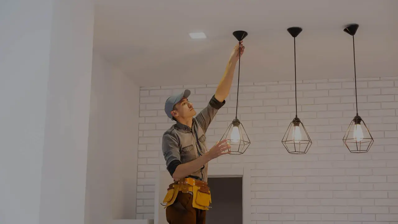 Fachowiec w pasie z narzędziami dokonuje montażu kinkietu, wiszącego obok innych lamp.
