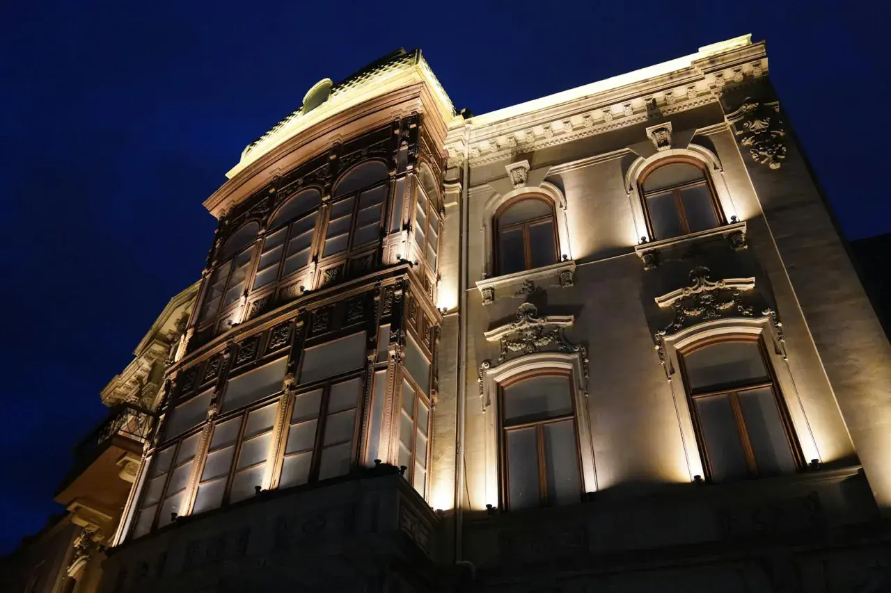 Elegancka kamienica nocą, oświetlona ciepłym blaskiem lamp miejskich. Widać bogate zdobienia fasady i duże, łukowe okna.