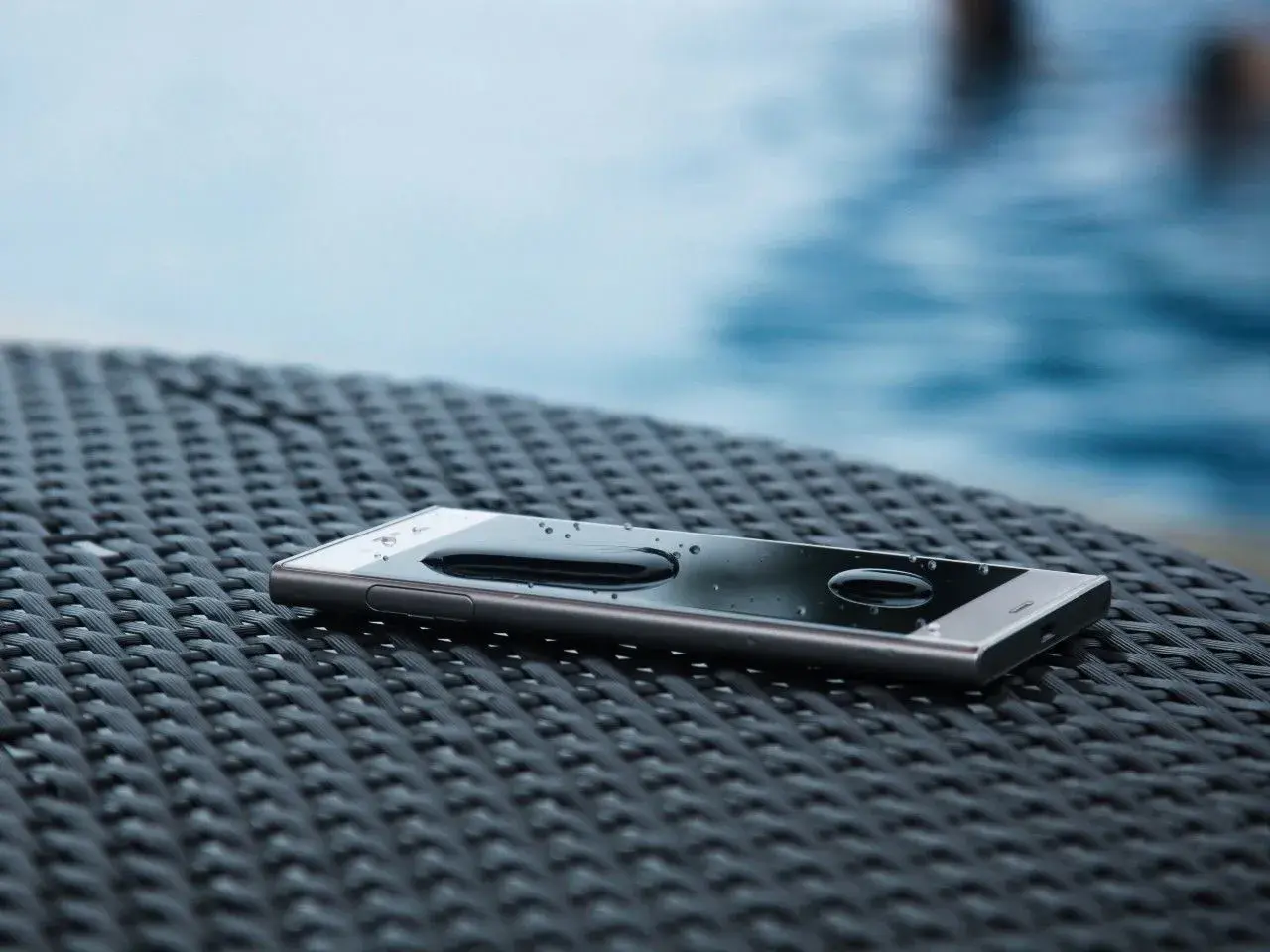Smartfon z kroplami wody na ekranie, leżący na plecionej powierzchni. To dowód, że ip68 co to znaczy: ochrona przed wodą i pyłem.