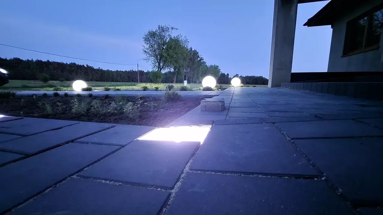 Nocny ogród rozświetlony kulistymi lampami i ledami w kostce brukowej. Ścieżka z ciemnej kostki brukowej prowadzi do domu.