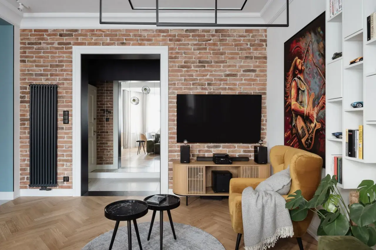 Nowoczesny salon z ceglaną ścianą, na której wisi telewizor. Oświetlenie ściany z kamienia podkreśla jej fakturę. Obok żółty fotel i obraz z gitarzystą.