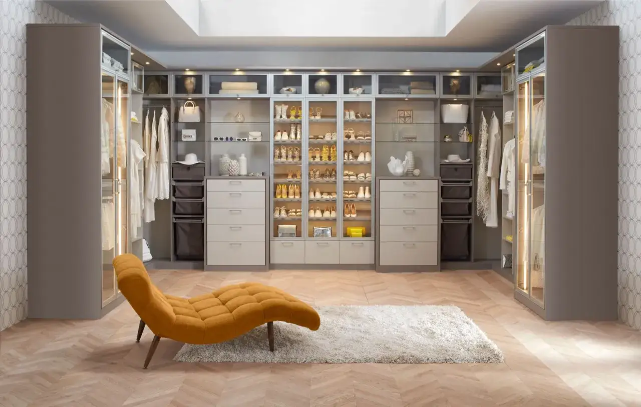 Elegancka garderoba z półkami pełnymi butów i ubrań, podkreślona dyskretnym oświetleniem. Pomieszczenie dopełnia designerski szezlong.