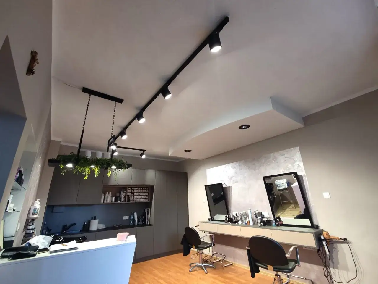 Nowoczesne oświetlenie salonu fryzjerskiego tworzy przytulną atmosferę. Reflektory na szynach i wiszące lampy z zielenią podkreślają styl wnętrza.
