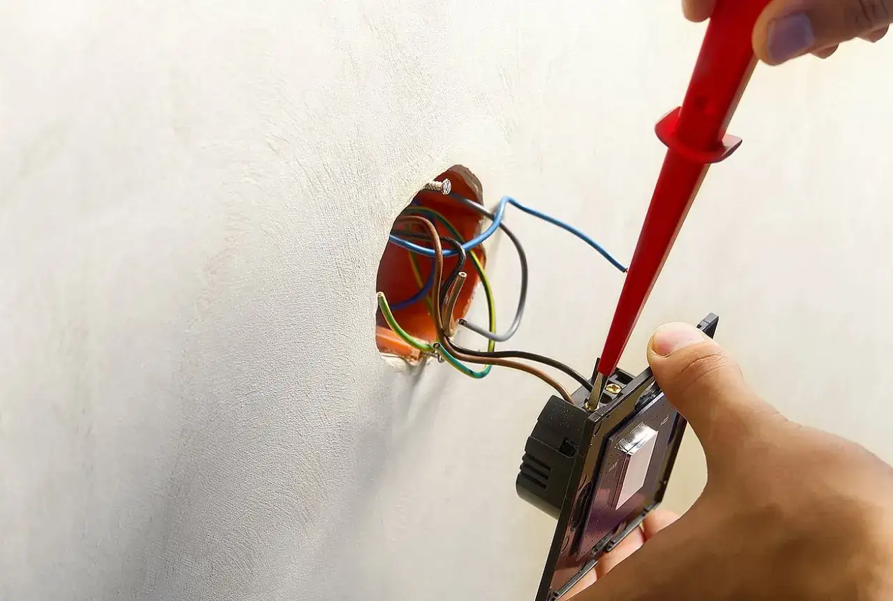 Instalator podłącza włącznik światła 2 kable, używając próbnika napięcia. Widać przewody w puszce instalacyjnej.