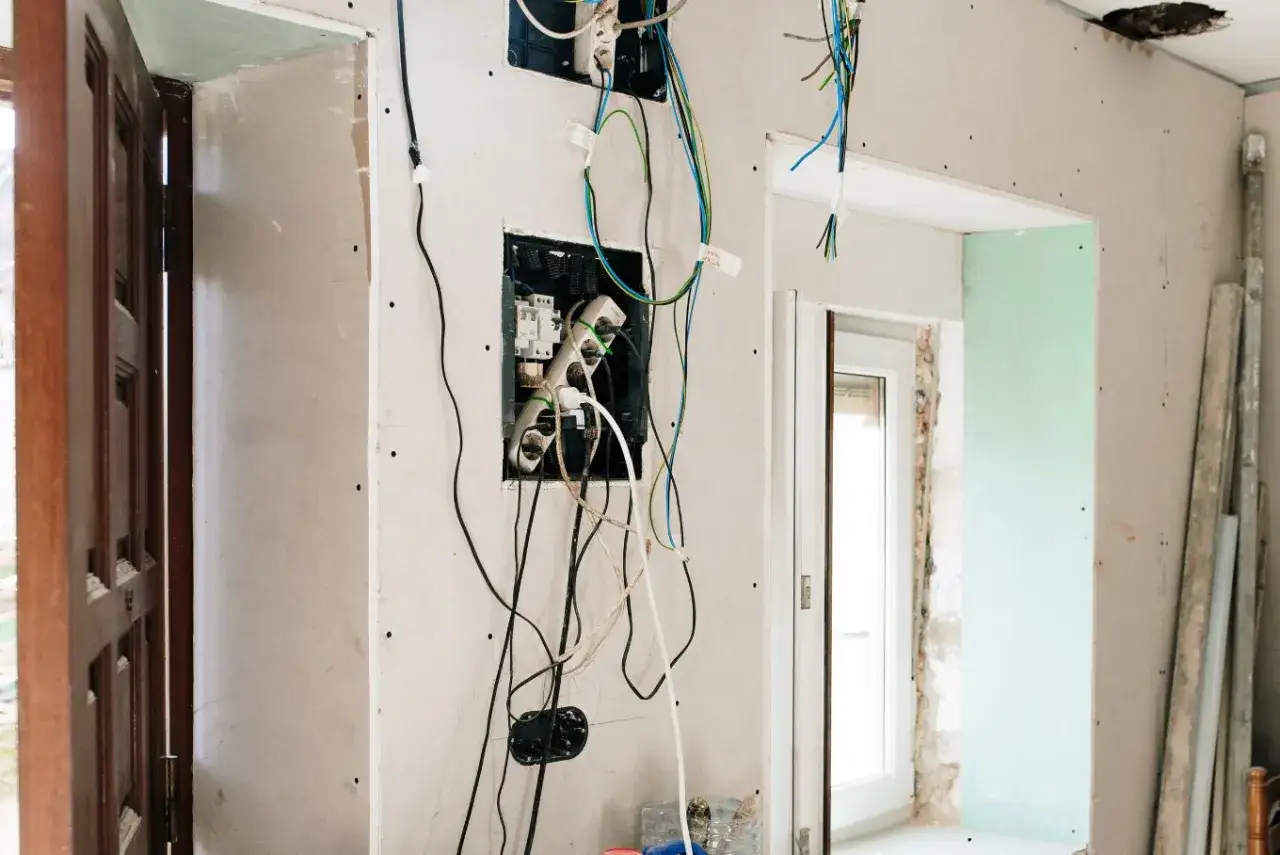 Widok na ścianę w trakcie remontu, z widoczną instalacją elektryczną i luźno zwisającymi przewodami. W ścianie widać otwory na rozdzielnicę i gniazdka, a obok drzwi i okno. To miejsce czeka na swoją kostkę do kabli.