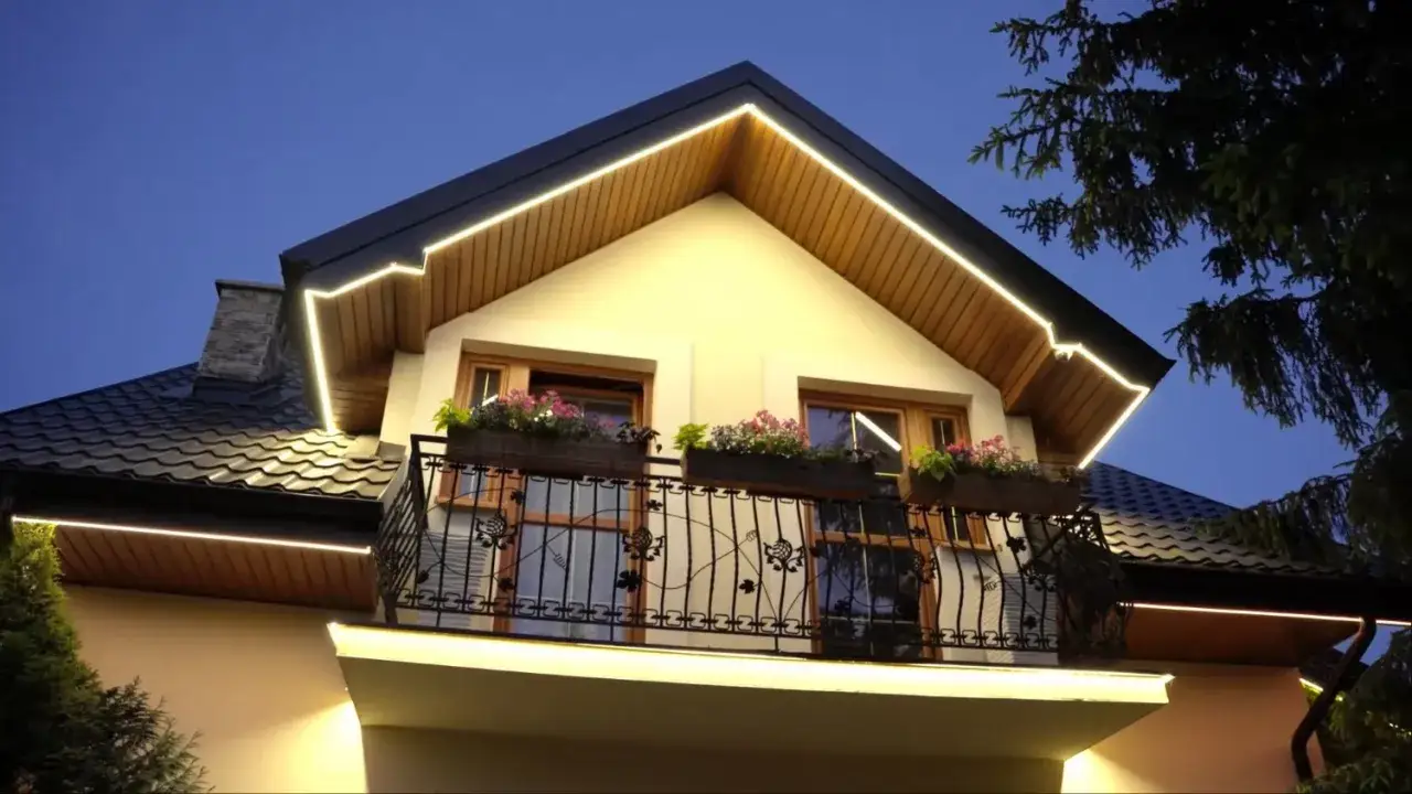 Elegancki dom nocą, podkreślony ciepłym światłem led w podbitce. Balkon z kwitnącymi kwiatami i ozdobną balustradą.